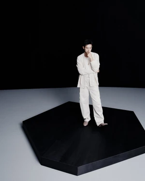Li Xian standing on a black hexagonal platform wearing cream-colored attire against a stark background