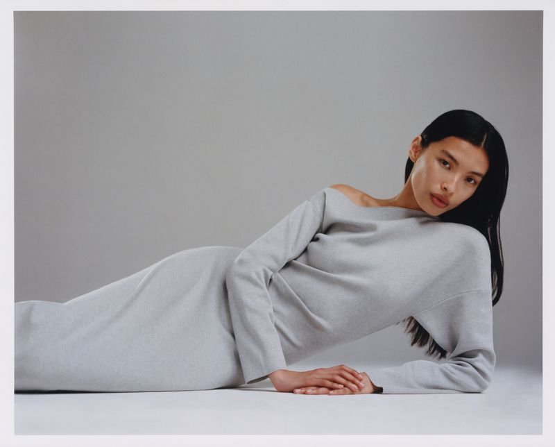 Woman lying down wearing a grey, long-sleeved dress, gazing directly at the camera in a studio setting with soft lighting.