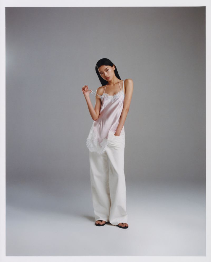 A slender Asian woman poses in a light pink lace-trimmed camisole and loose white pants against a neutral grey background.