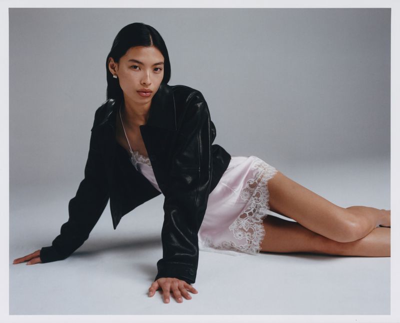 An adult woman poses reclining in a pink slip dress and black jacket against a neutral studio backdrop, fashion, and portrait photography.
