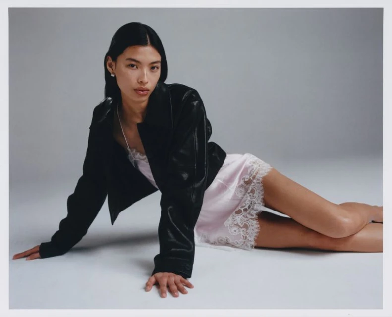 An adult woman poses reclining in a pink slip dress and black jacket against a neutral studio backdrop, fashion, and portrait photography.