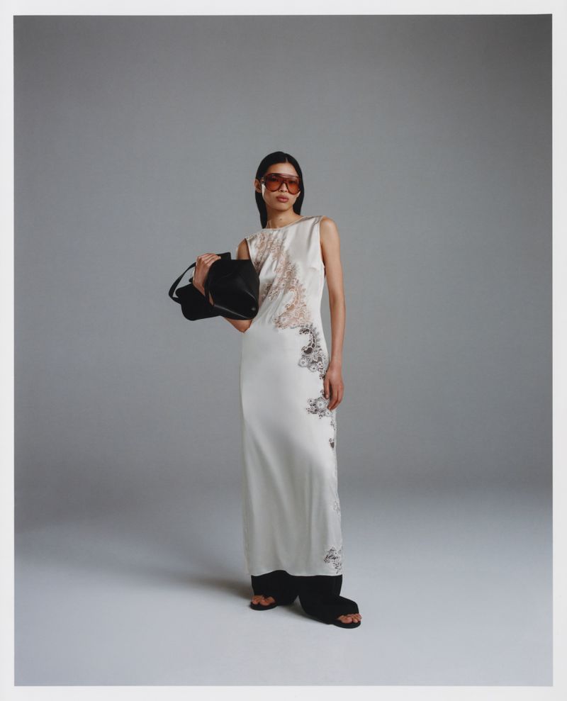 A slender Asian woman in a floor-length patterned dress poses in a studio with a black handbag and amber-tinted glasses, styled and graceful.