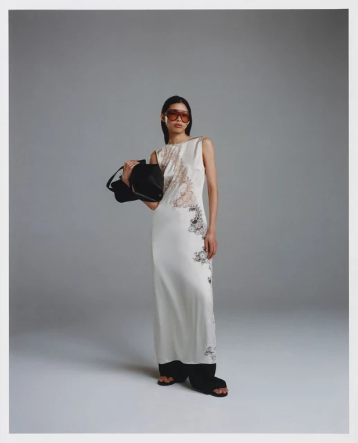 A slender Asian woman in a floor-length patterned dress poses in a studio with a black handbag and amber-tinted glasses, styled and graceful.
