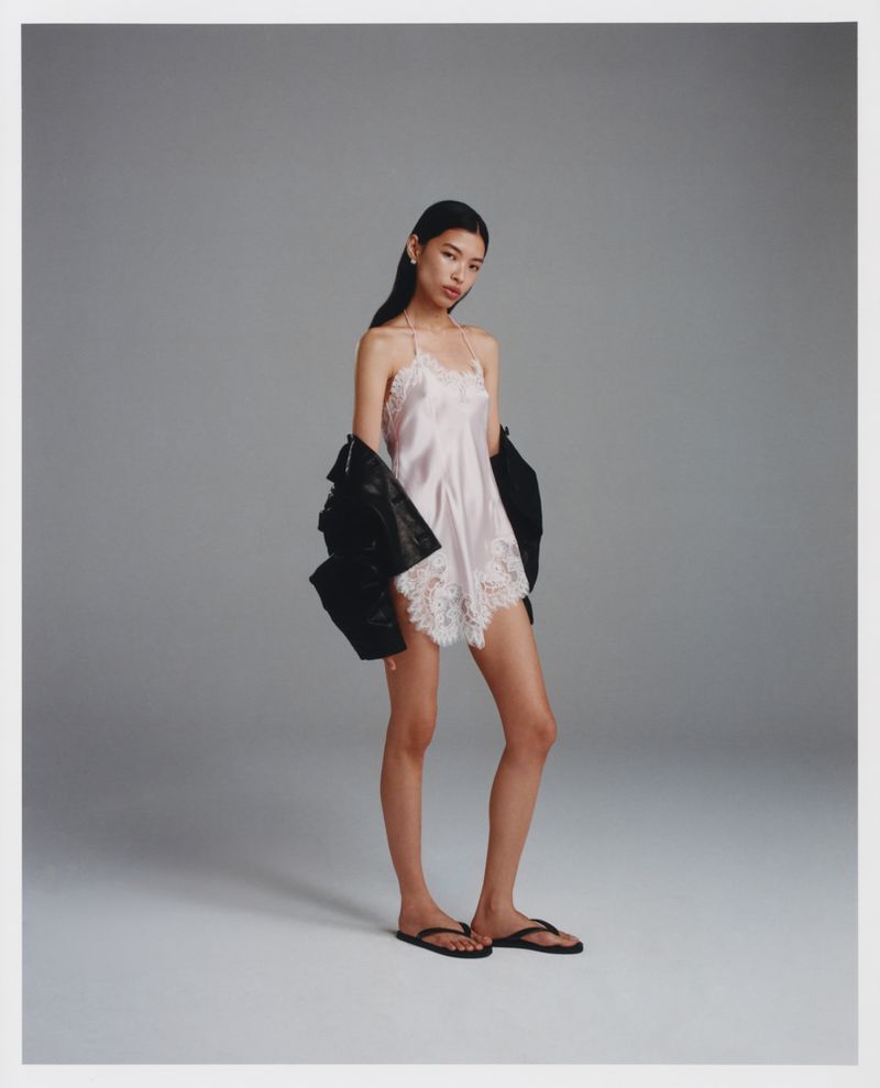 Full-length shot of a woman in a slip dress and flip flops in a studio, wearing a black leather jacket slung over her shoulder.