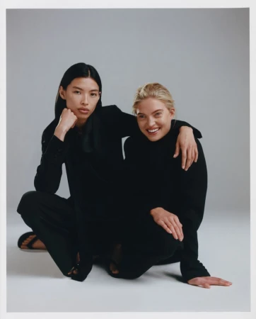 Two female models in black outfits posing together against a plain grey studio backdrop for Revolve.
