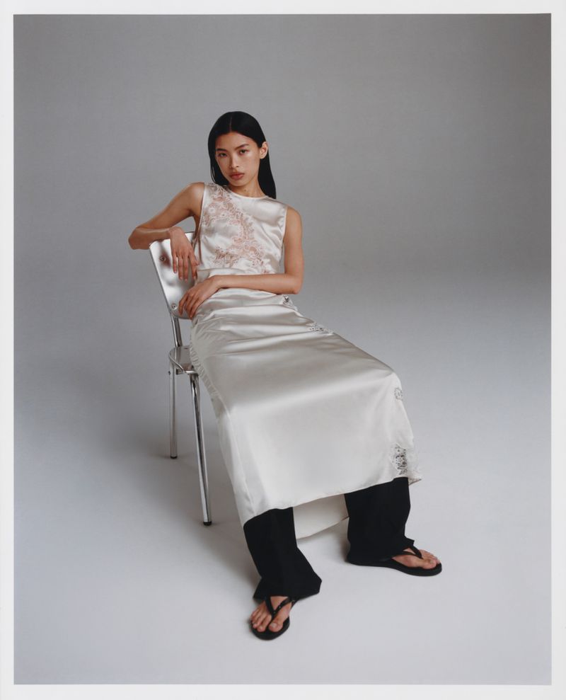 A slender woman with long black hair is seated on a chrome chair. She wears a cream-colored, satin two-piece dress with black sandals in studio setting.