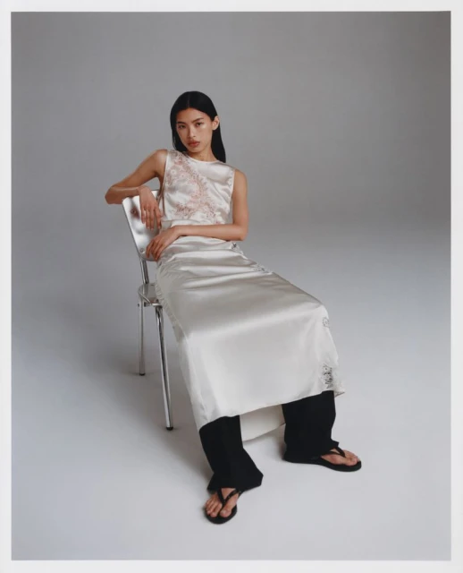 A slender woman with long black hair is seated on a chrome chair. She wears a cream-colored, satin two-piece dress with black sandals in studio setting.