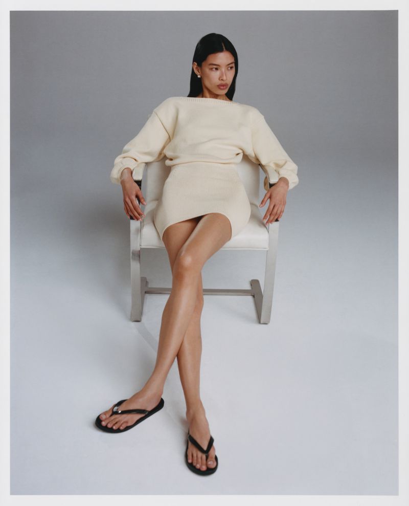 Elegant Asian woman in cream knitwear and black flip-flops seated in a white chair against a neutral studio backdrop. Fashion shot.