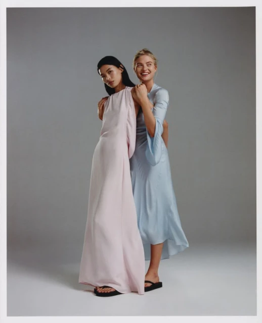Two adult female models in light-colored dresses, one pink, one light blue, posing together in a studio with a grey background.