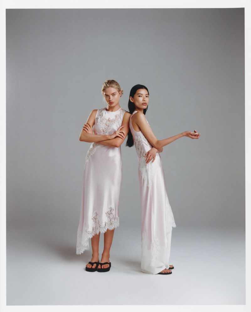 Two fashion models in pink gowns and black sandals pose back-to-back in a photo studio with a soft, neutral background, full shot.