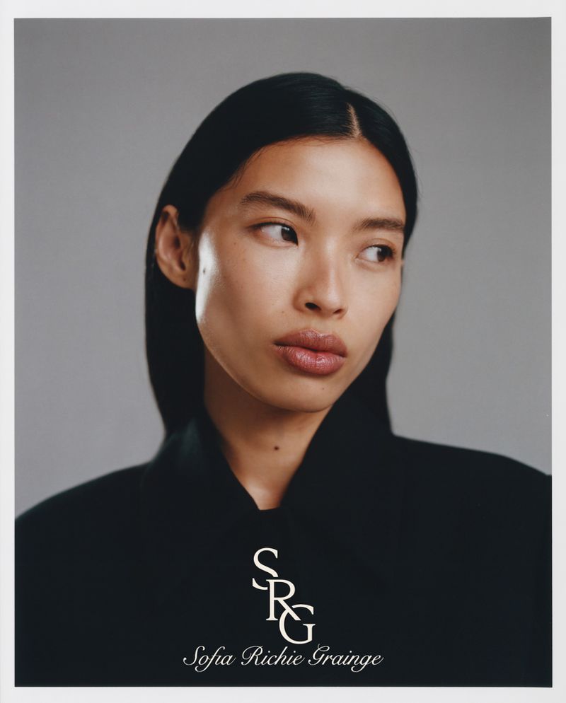Close-up portrait of a woman with sleek dark hair and natural makeup against a plain grey studio backdrop with white border.