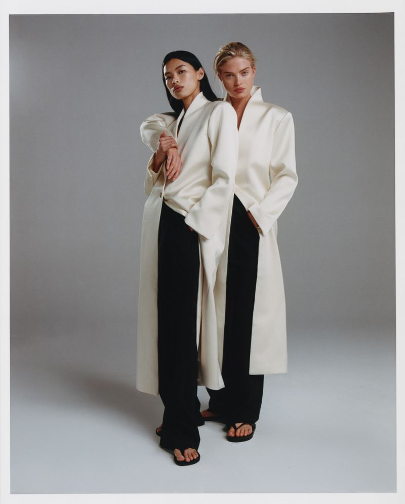 Two models standing together wearing matching white coats and black trousers against a neutral grey studio backdrop.