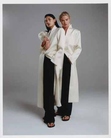 Two models standing together wearing matching white coats and black trousers against a neutral grey studio backdrop.