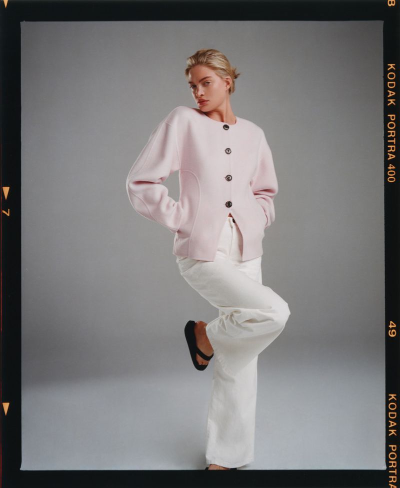 Model wearing a pink buttoned cardigan and white pants posing against a grey seamless background.