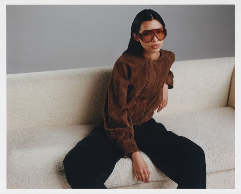 Model wearing brown jacket and tinted sunglasses seated on a cream textured sofa