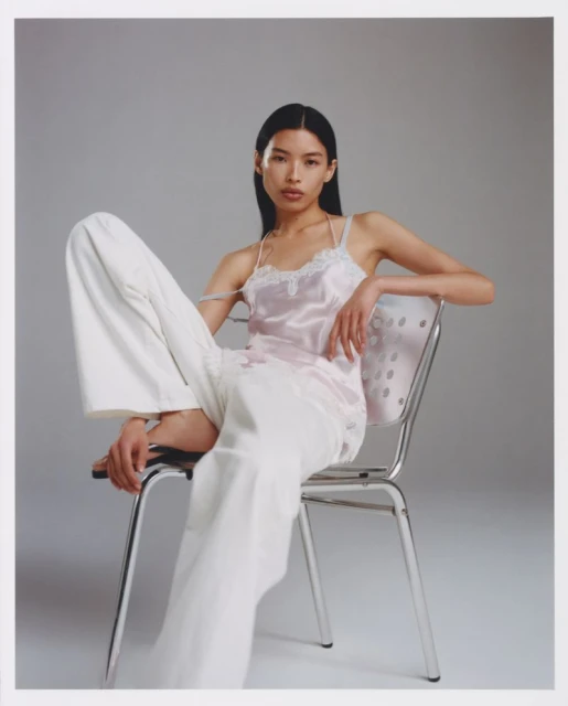 Adult woman with long dark hair sits on metal chair wearing a satin camisole and white pants against a neutral backdrop in a fashion shot.