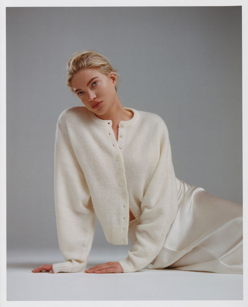 Blonde woman in a white cardigan and silk skirt poses in a studio with soft lighting, creating a minimalist fashion portrait.