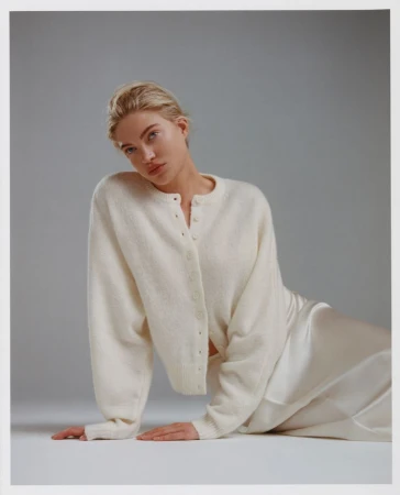 Blonde woman in a white cardigan and silk skirt poses in a studio with soft lighting, creating a minimalist fashion portrait.