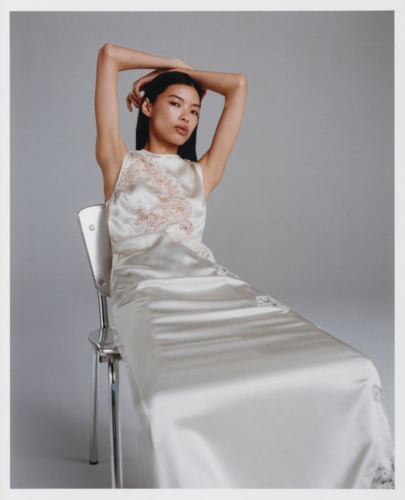 Woman in a satin dress sitting on a metal chair in a studio with a neutral background, arms raised behind her head.