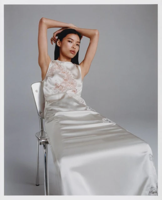 Woman in a satin dress sitting on a metal chair in a studio with a neutral background, arms raised behind her head.