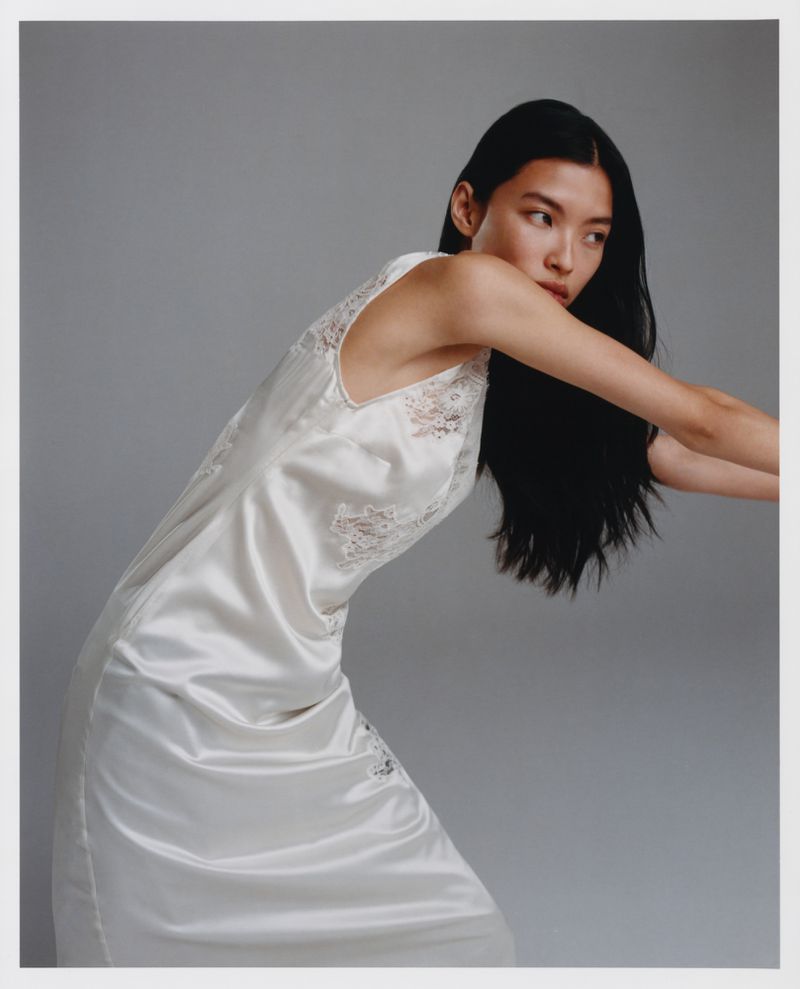 Elegant Asian woman in a white satin and lace dress posing against a gray backdrop in a fashion forward pose with dark hair.
