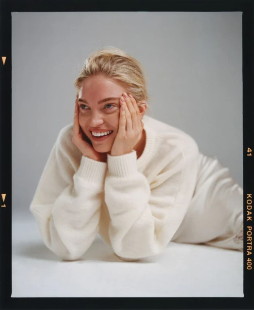 Smiling blond woman in a cream sweater rests her chin on her hands in a studio portrait with diffused lighting and a clean backdrop.