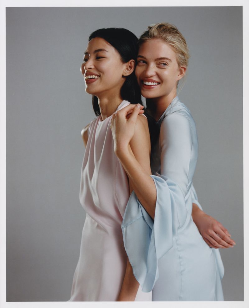 Two smiling women in pastel dresses pose indoors against a grey background, sharing an embrace, exemplifying fashion and friendship.