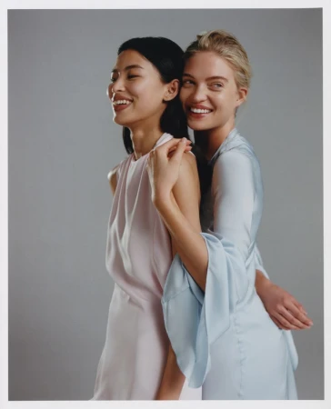Two smiling women in pastel dresses pose indoors against a grey background, sharing an embrace, exemplifying fashion and friendship.