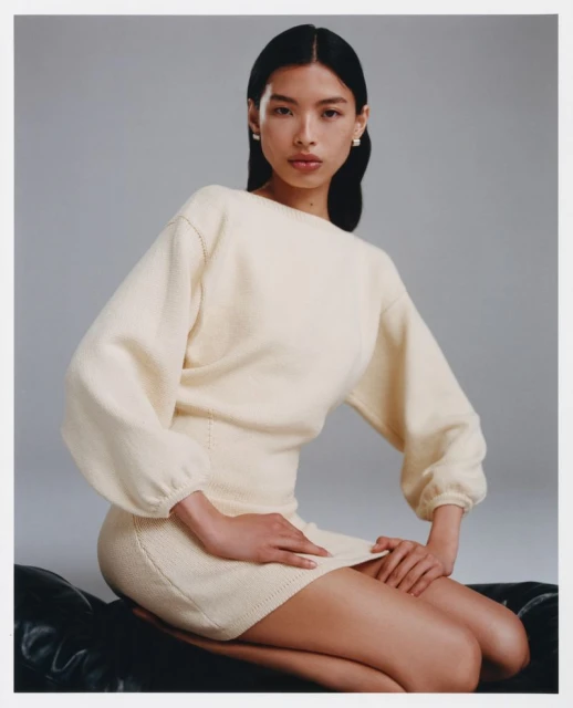 Portrait of an Asian woman in an off-white sweater dress seated indoors on a dark surface with a neutral gray background.