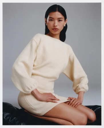 Portrait of an Asian woman in an off-white sweater dress seated indoors on a dark surface with a neutral gray background.