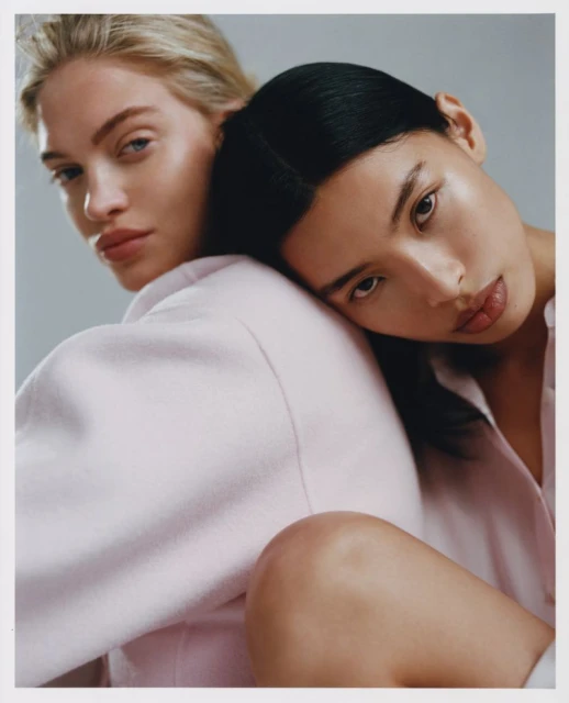 Two women with dark and blonde hair pose closely indoors. They wear light pink shirts and neutral makeup against a grey backdrop.