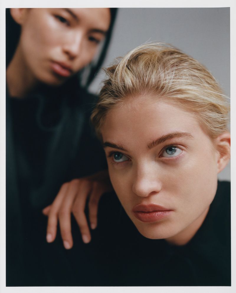 Close-up portrait of two women with fair skin and neutral expressions, one with blonde hair and one with dark hair, in front of a grey background.
