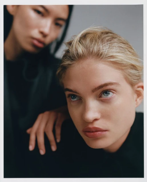 Close-up portrait of two women with fair skin and neutral expressions, one with blonde hair and one with dark hair, in front of a grey background.