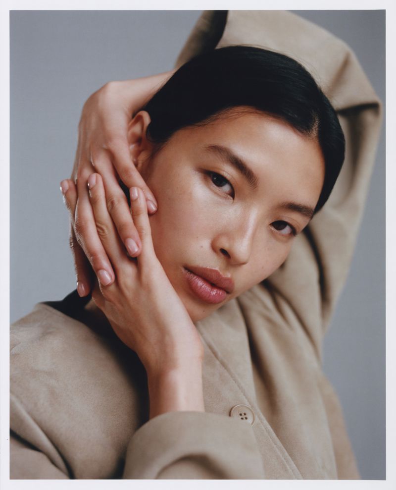 Close-up portrait of an Asian woman with hands near her face. She is wearing a beige jacket against a light gray background.