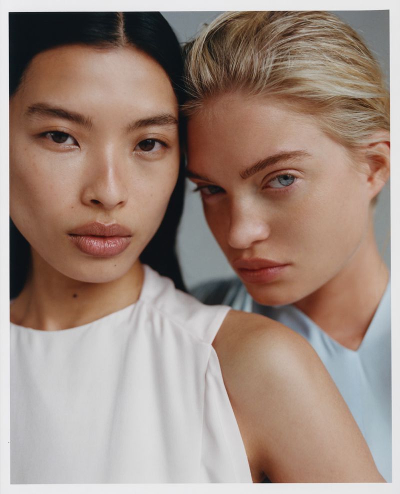 Two adult women pose in a close-up shot with neutral facial expressions, wearing light-colored tops against a light grey backdrop.