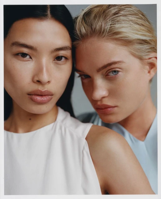 Two adult women pose in a close-up shot with neutral facial expressions, wearing light-colored tops against a light grey backdrop.