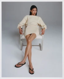 Woman in cream knit dress seated on a white chair against a plain grey backdrop