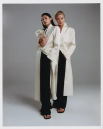 Two models standing together wearing matching white coats and black trousers against a neutral grey studio backdrop.
