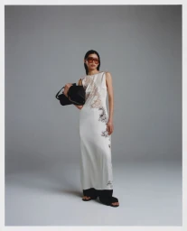Model in white patterned dress holding black handbag wearing sunglasses against a grey studio backdrop