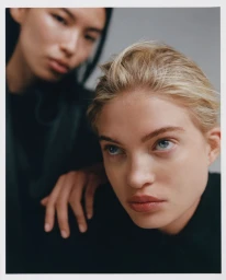 Portrait of a blonde woman with blue eyes in the foreground and a dark-haired woman behind her against a grey background.