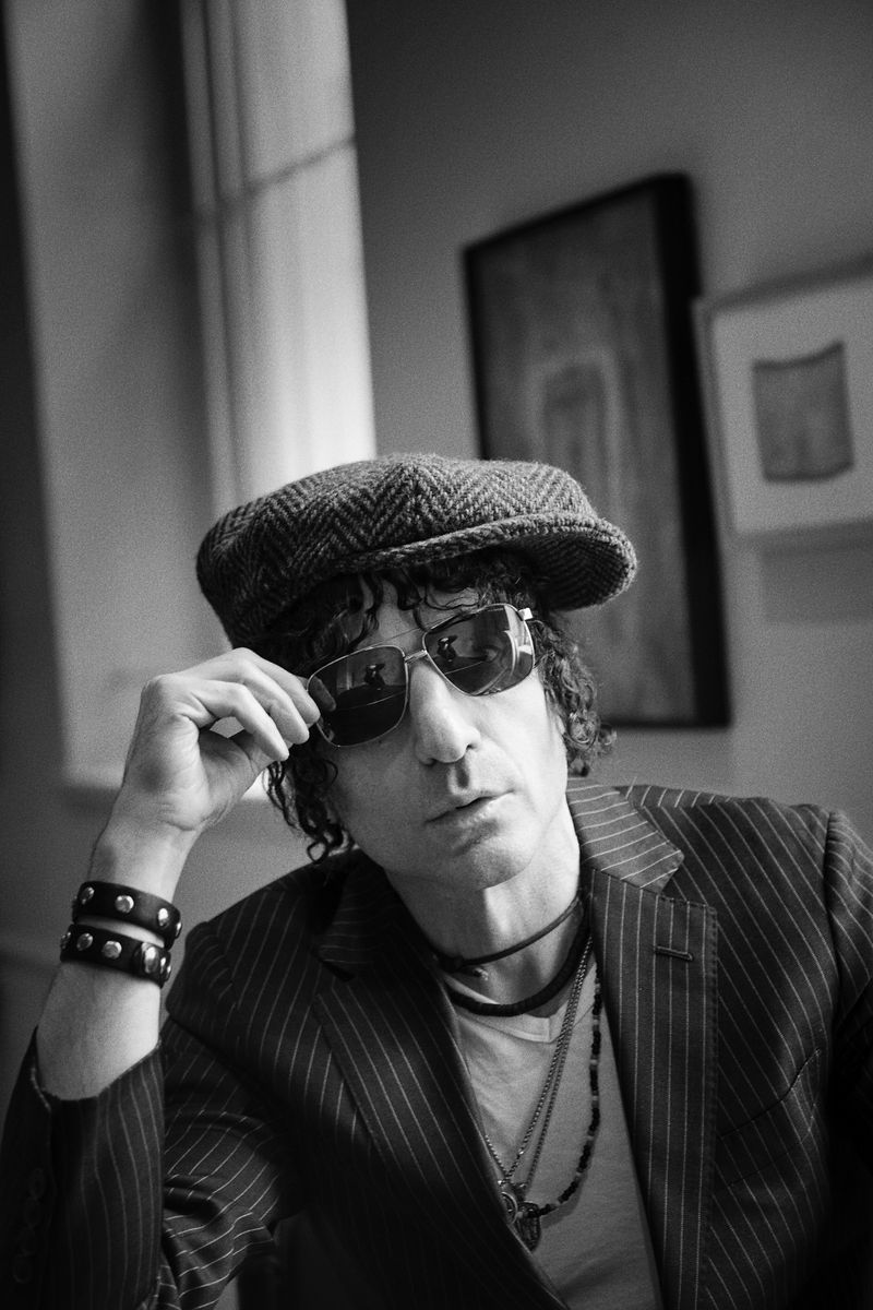 Black and white portrait of a stylish adult male with sunglasses and a cap, wearing a pinstripe jacket and jewelry indoors.
