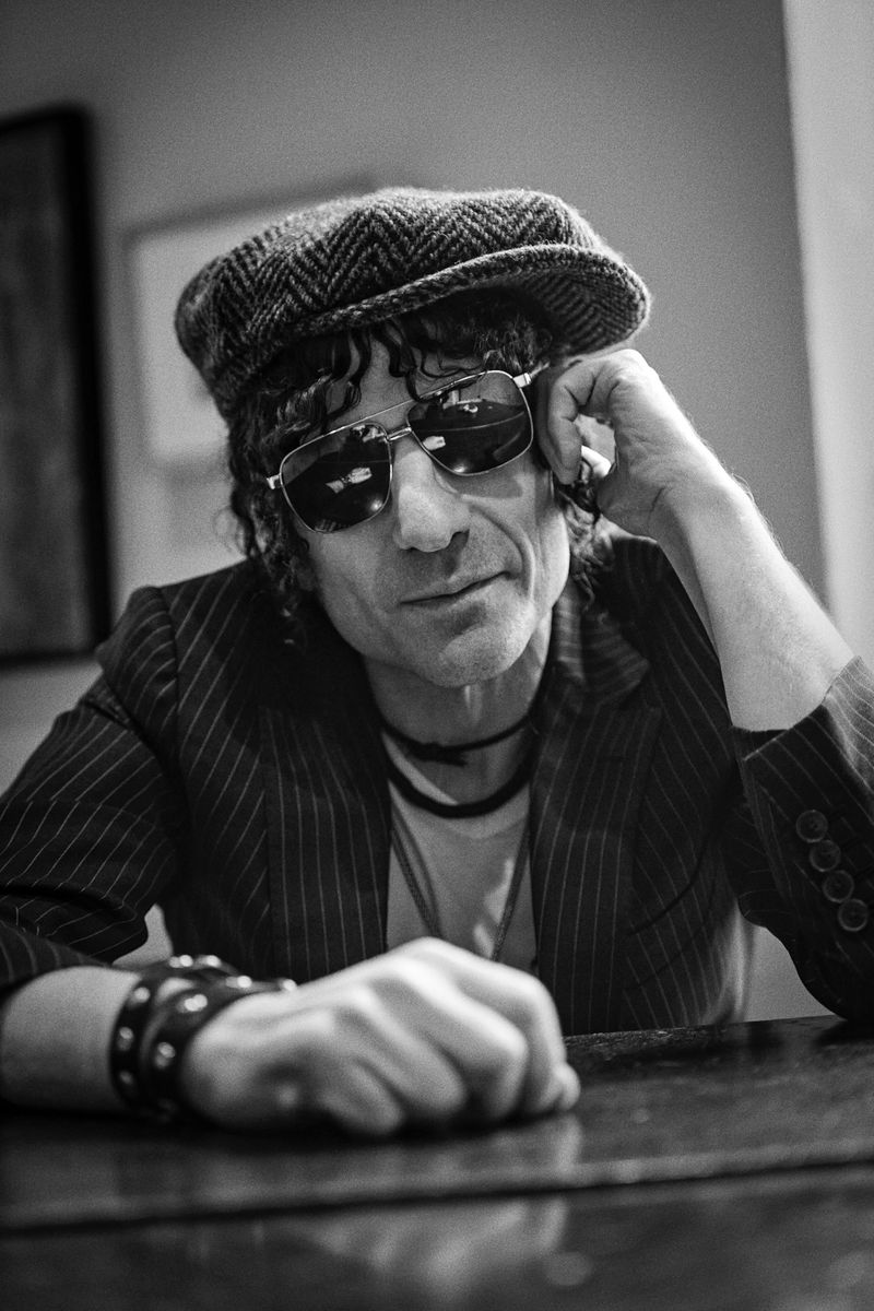 Monochrome portrait of a seated man in a pinstripe jacket, newsboy cap, and sunglasses, resting his hand on a dark table.