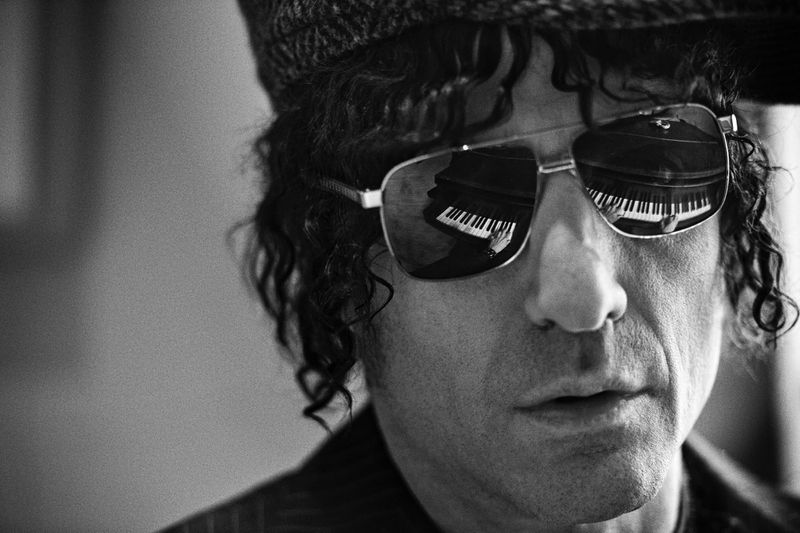 Black and white portrait of adult man with curly hair wearing aviator sunglasses that reflect a piano in an indoor setting.