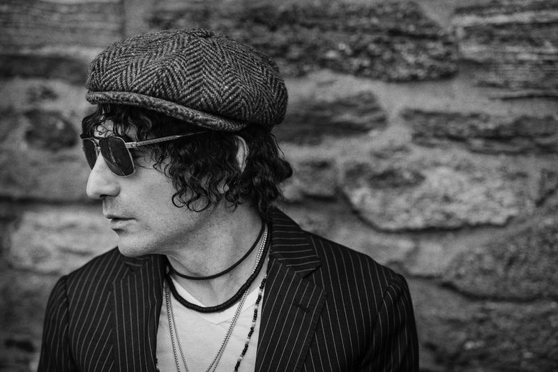 Black and white portrait of a man with curly hair wearing a cap, sunglasses, striped blazer standing against a stone wall.