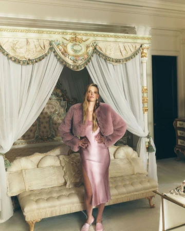 Elegant woman in pink dress and faux fur jacket poses in luxury bedroom with four-poster bed, evoking fashion and interior design.