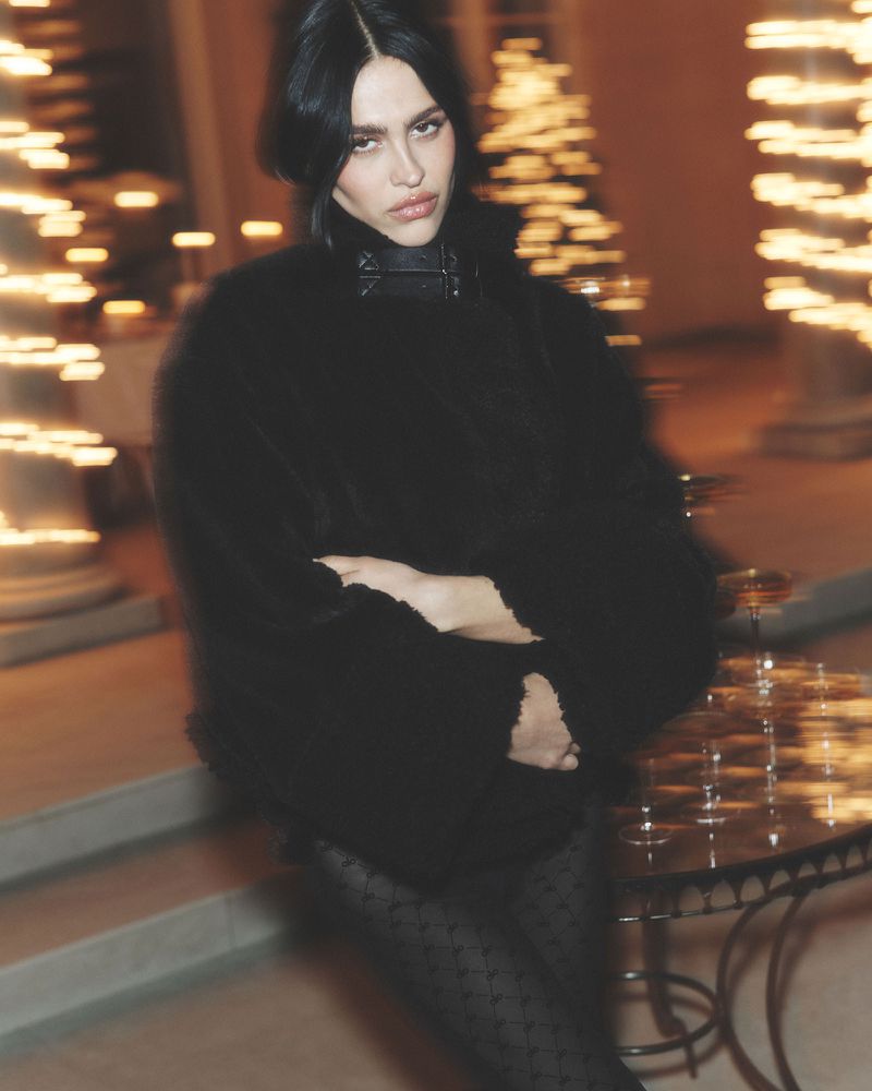 Portrait of a fashionable young adult woman with dark hair and a black coat posing near a table with glassware and blurred lights.