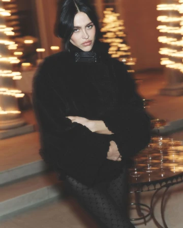 Portrait of a fashionable young adult woman with dark hair and a black coat posing near a table with glassware and blurred lights.