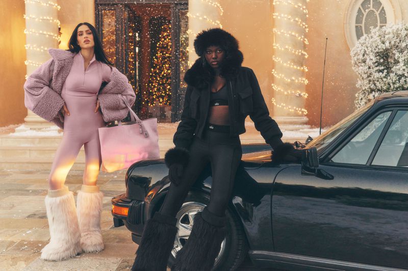 Two women pose in Alo Yoga winter apparel beside a black car in front of a house decorated with holiday lights and snowy landscape.