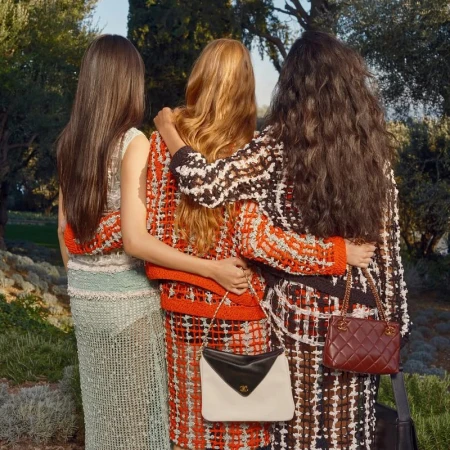 Three women stand together in a sunlit outdoor setting, showcasing knitwear and purses in a fashion editorial. The scene has soft natural light.