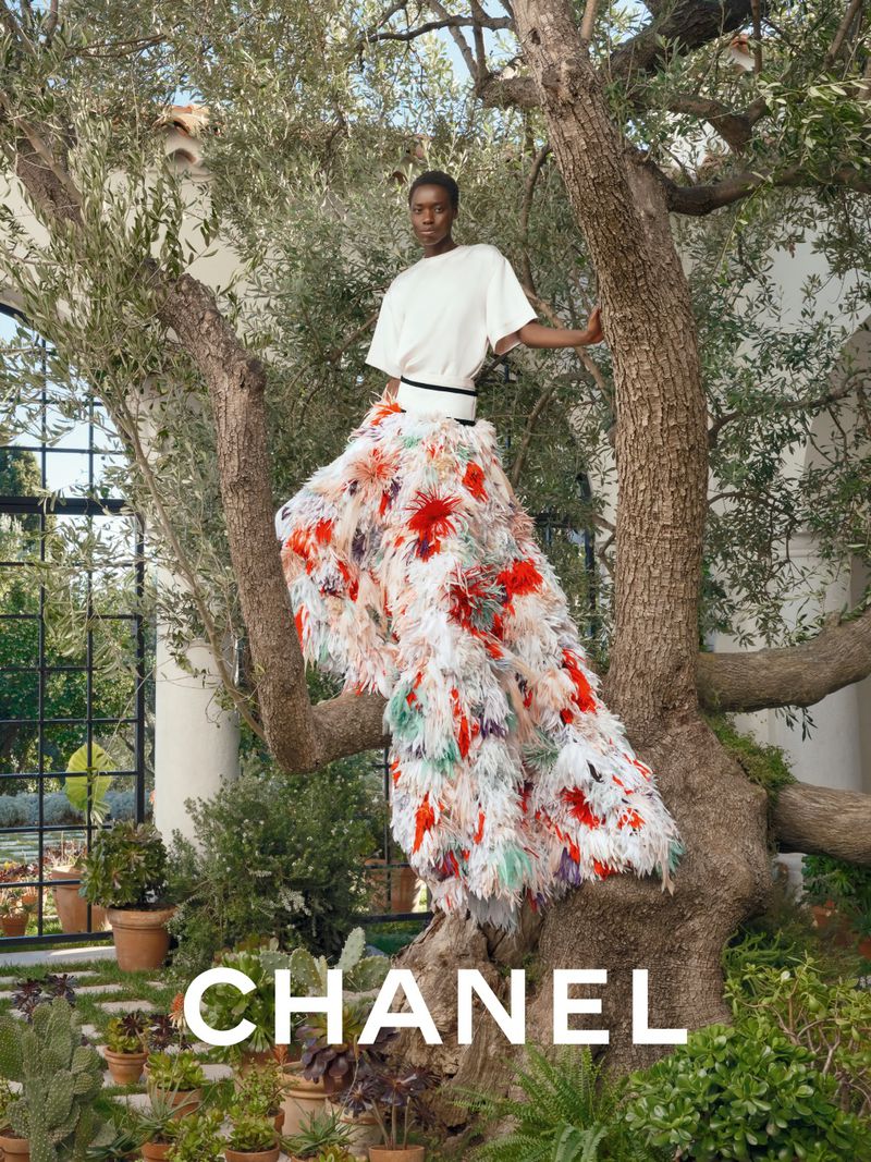 Fashion shot of an African woman posing in a floral feather skirt and white top, standing on a tree branch outdoors with Chanel logo.